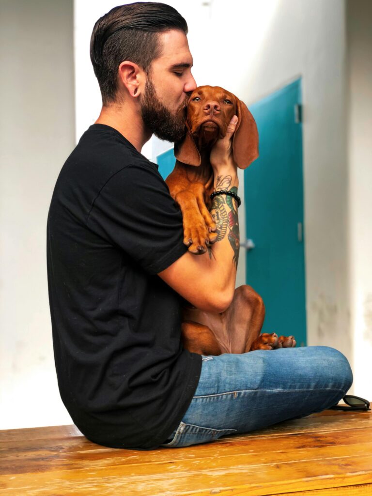 A bearded man sits kissing his dachshund puppy in a casual setting, showing love and companionship.